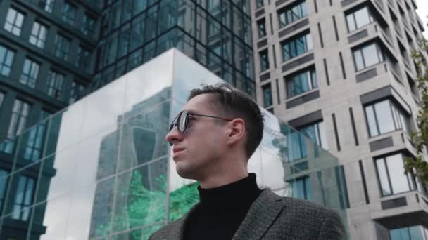 Retrato del hombre de negocios atento que usa traje y gafas de sol mirando hacia fuera mientras que está parado cerca del edificio de oficinas moderno. Joven empresario masculino mirando alrededor en el centro de la ciudad. Parece exitoso.. — Vídeo de stock