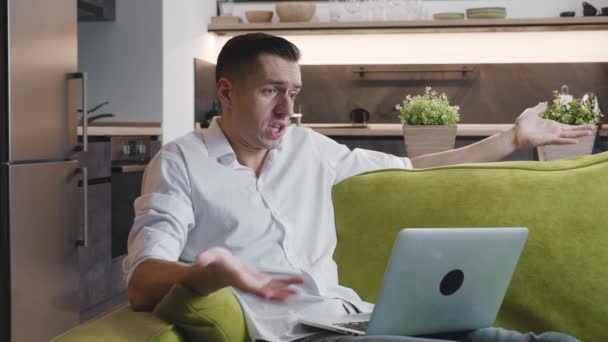 Angry man having an online video call via laptop computer with a friend shows resentment and aggressive emotions to the opponent while lying on sofa at home. — Stock Video