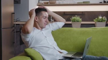 Neşeli genç erkek işadamı evdeki rahat bir kanepede yatarken dizüstü bilgisayarda çalışmaktan zevk alıyor. Erkek bir serbest çalışan evden uzakta çalışıyor..