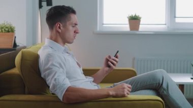 Sinirli genç adam elinde akıllı telefon tutuyor. Kötü mesajdan bıkmış, evdeki kanepede oturuyor. Kızgın erkek cep telefonunda uygulama kullanıyor, yumruğunu kanepeye vuruyor ve bağırıyor. Saldırganlık göstergesi.