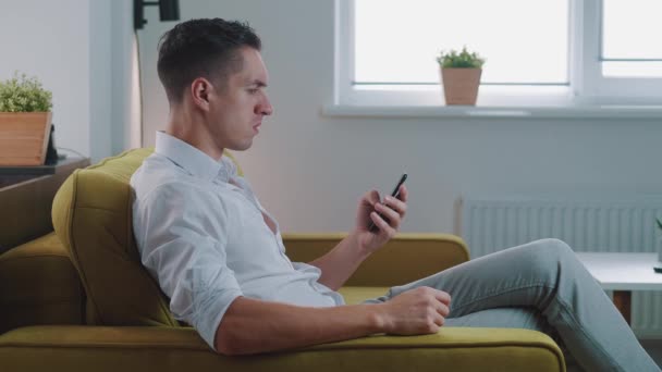 Annoyed young man holding smartphone frustrated by bad message, sitting on sofa at home. Angry male uses application on his mobile phone, bangs his fist on the sofa and shouts. Display of aggression. — Stock Video