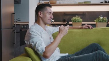 Evdeki kanepede dinlenirken akıllı telefon kullanan gülümseyen genç adam. Oturma odasındaki rahat koltukta dinlenirken cep telefonu cihazında mesajlaşan neşeli erkek bandı..