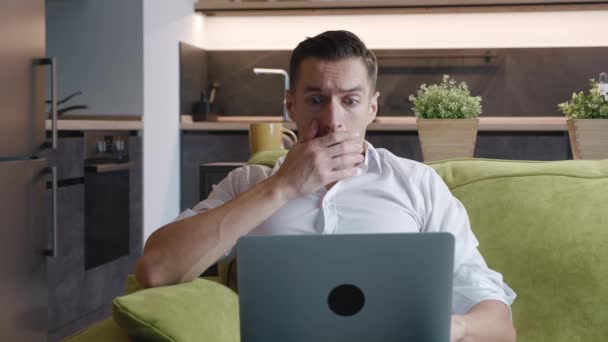 怖いビジネスマンは、問題を心配してノートパソコンの画面を見てストレスを感じる悪いオンラインニュースを読む。自宅でソファに座って、コンピュータ上の恐ろしいニュースで彼の口をカバー恐怖. — ストック動画