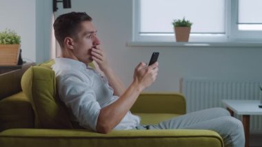 Smartphone 'daki şok olmuş adam ev oturma odasındaki kanepede otururken ağzını korkuyla kapatıyor. Nahoş bir şekilde şaşırmış ve korkmuştu. Erkek, gördüğü şey karşısında şoke olur..