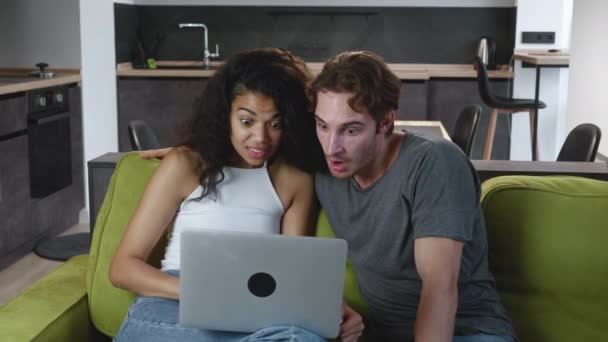 Erstauntes gemischtes Paar liest gute Nachrichten auf Laptop-Computer, junge Mann und Frau sind angenehm überrascht und zeigen WOW Emotionen, während sie es sich zu Hause auf der Couch gemütlich machen. Aufgeregt. — Stockvideo