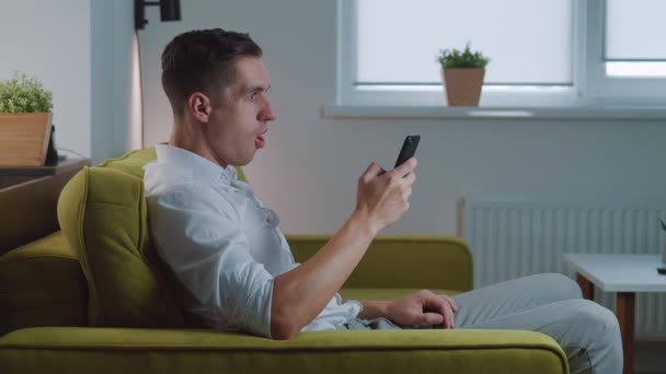 Aufgeregter überglücklicher Sieger, der den Erfolg feiert, auf das Smartphone schaut und zu Hause auf der Couch eine Geste macht. Mobiles Siegeskonzept — Stockvideo