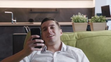 Mutlu adam işten sonra kanepeye düşüyor ve evde tatilin tadını çıkarırken akıllı telefon kullanıyor. Genç gülen işadamı uygulamayı evdeki kanepede dinlenirken cep telefonuyla kullanıyor..