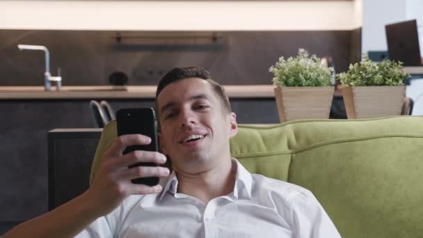 El hombre feliz se cae en el sofá después del trabajo y utiliza el teléfono inteligente mientras disfruta de sus vacaciones en casa. Joven hombre de negocios sonriente utiliza la aplicación en el teléfono móvil mientras se relaja en el sofá en casa. — Vídeos de Stock