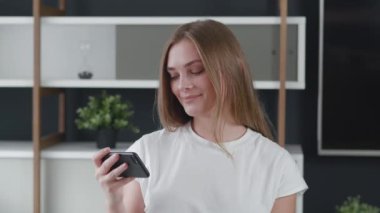 Güzel, genç, gülümseyen bir kadın sosyal ağlar için akıllı telefon kullanıyor, cep telefonu videoları izliyor, arkadaşlarıyla iletişim kuruyor..