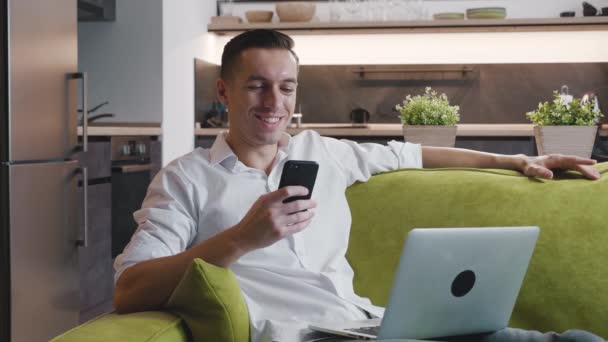 Giovane imprenditore maschio felice utilizza smartphone mentre si lavora in un computer portatile seduto a casa sul divano. — Video Stock