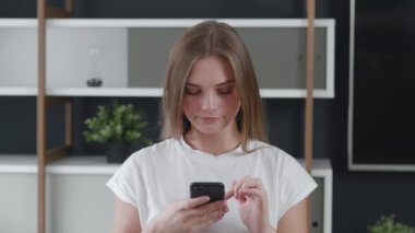 Ofiste akıllı telefon kullanan gülümseyen genç bayan öğrenci, modern ofis arka planında sosyal medya için cep telefonu kullanan mutlu iş kadını. İnterneti tarıyor, arkadaşlarıyla iletişim kuruyor..