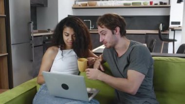 Mutlu çok ırklı genç çift konuşuyor ve birlikte dizüstü bilgisayar kullanıyorlar. Oturma odasındaki kanepede oturup video izliyorlar. Beyaz adam ve Afro-Amerikan kadın evdeki bilgisayara bakıyor..
