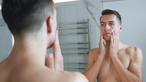 鏡を見て顔にクリームを塗る若い男。ハンサムな若い男性は自宅のバスルームで彼の顔に保湿剤を適用します。スキンケアのコンセプト. — ストック動画
