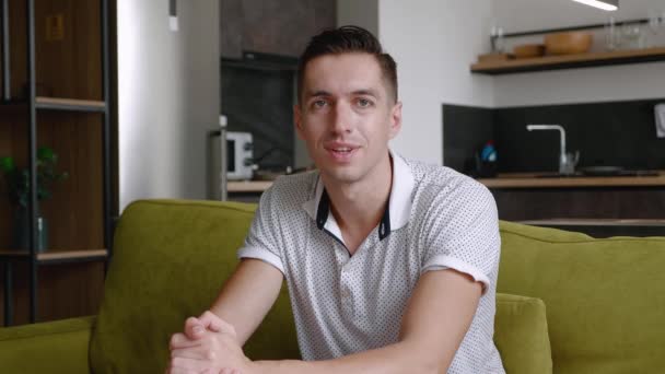 Retrato del hombre sonriente descansando mientras está sentado en el sofá en casa. Un joven macho disfruta del estilo de vida mirando a la cámara mientras está sentado en un acogedor sofá en su apartamento. — Vídeos de Stock