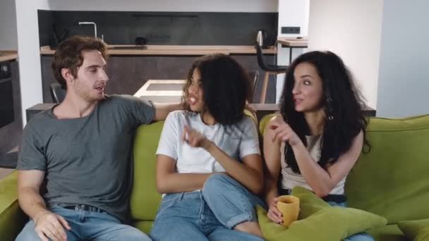 Группа из трех друзей один мужчина и две женщины разговаривают, смеясь вместе, сидя на диване. Молодые люди смешанной расы устраивают вечеринку дома — стоковое видео