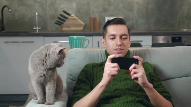 Νεαρός άνδρας με πουλόβερ που παίζει παιχνίδια εφαρμογής στο smartphone του ενώ ξεκουράζεται στον καναπέ στο άνετο σπίτι με οικιακή γάτα. — Αρχείο Βίντεο