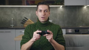 Oturma odasındaki kanepede otururken video oyunu oynayan heyecanlı adam oyuncuyu tutuyor. Genç, gülümseyen bir erkek video oyununda sevinçten havalara uçuyor.