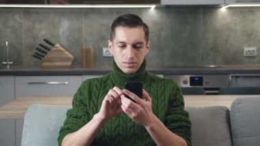 Mutlu genç adam cep telefonundan iyi haberler alıyor. Heyecanlı erkek kazanan mobil zafer kavramını kutluyor akıllı telefona bakıyor ve evdeki kanepede otururken evet jestleri yapıyor..