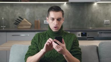 Evdeki kanepede otururken akıllı telefonuyla haber okuyan şok olmuş bir adam. Hoş olmayan bir şekilde şaşırmış ve korkmuş genç bir erkek ev oturma odasındaki kanepede ağzını korkuyla kapatıyor. Cep telefonu kullanan adam..