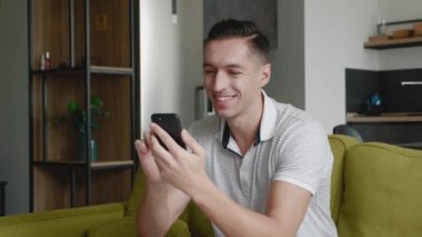 Modern apartmandaki akıllı telefon tebessümü kullanarak kanepede oturan neşeli genç adama yakın çekim teknolojisi mesajları yolluyorum.