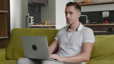 Evdeki koltukta dizüstü bilgisayar kullanan serbest çalışan genç bir adam. Dairede bilgisayarla uzaktan çalışan bir erkeğin portresi.