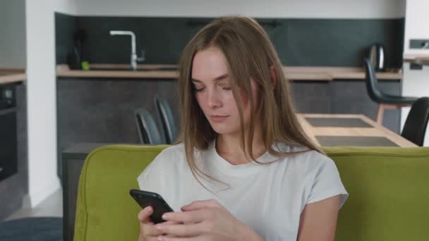 Terkejut dan khawatir wanita muda membaca berita buruk di telepon pintarnya sambil duduk di sofa di rumah. — Stok Video