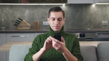 Yakışıklı beyaz adam rahat evinde kanepede oturuyor ve akıllı telefon kullanıyor sosyal medyada geziniyor, videolar izliyor ve gülümsüyor..