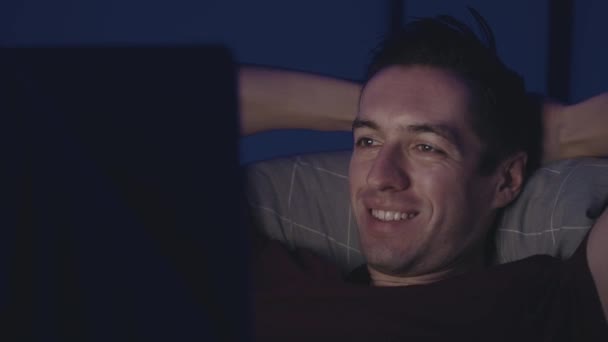 夜にベッドで休んでいる間、ノートパソコンで映画を見て笑っている若い男。夜間に自宅の寝室に横たわっている間、ラップトップコンピュータを使用して男性. — ストック動画