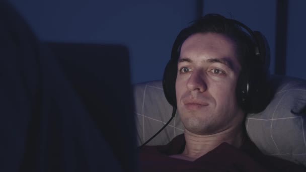 Kulaklıklı genç adam geceleri yatağında dinlenirken dizüstü bilgisayarında film izliyor. Erkek, geceleri yatak odasında yatarken dizüstü bilgisayar kullanıyor.. — Stok video