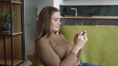 Evdeki kanepede oturan ve akıllı telefondan oyun oynayan genç ve güzel bir kadın.