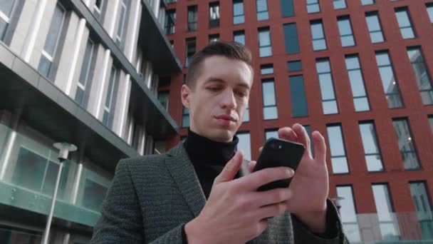 Retrato de homem sorridente empresário ficar na rua centro da cidade e usando smartphone. Jovem empreendedor de terno usando telefone celular ao ar livre no moderno edifício de fundo. — Vídeo de Stock