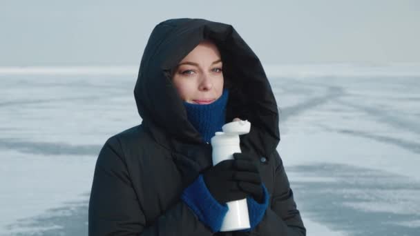 Young woman in warm down jacket with a hood, drinks hot drink from thermos in cold winter on arctic background. Portrait of a female warming herself with hot tea on background of frozen lake or river — Stock Video