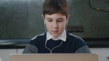 Evde ders çalışırken dizüstü bilgisayar kullanan beyaz bir çocuğun portresi. Kulaklıklı çocuk oturma odasında otururken bilgisayarda çalışıyor.