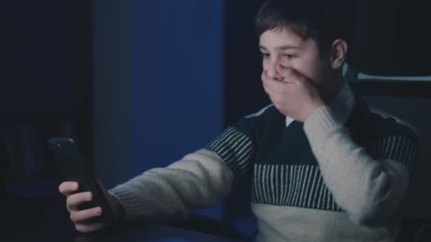 Chico asustado y sorprendido mira la pantalla del teléfono inteligente, cubre la boca con la mano en la noche en casa. Niño colegial asustado de malas noticias, leer terrible mensaje en el teléfono móvil sentado en la habitación. Viendo vídeo de terror — Vídeos de Stock