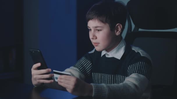 12-13 anos de idade menino compras on-line com cartão de crédito e smartphone, enquanto sentado em casa tarde da noite. Kid estudante com telefone celular usando sistemas de pagamento on-line. Banca online. — Vídeo de Stock