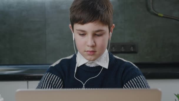 Retrato de adolescente caucasiano usando laptop enquanto estudava em casa. Menino escolar em fones de ouvido trabalhando no computador enquanto sentado na sala de estar — Vídeo de Stock