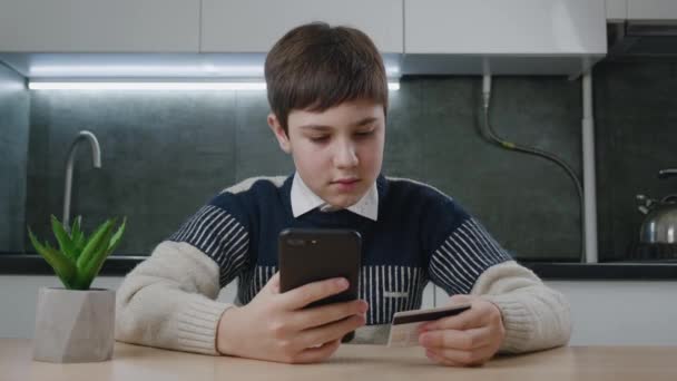 12-13 anos de idade menino sentado na mesa da cozinha e compras on-line com cartão de crédito e smartphone enquanto sentado em casa. Kid estudante com telefone celular usando sistemas de pagamento on-line. Banca online — Vídeo de Stock