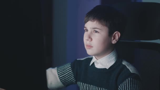 12-13 anos de idade menino usa computador à noite em casa. Aprendizagem infantil online. Criança pré-adolescente assistindo vídeo e usando aplicativos de computador durante a educação online. Monitore a reflexão da luz em seu rosto — Vídeo de Stock