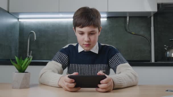 Portré jóképű fiúról, aki videojátékot játszik okostelefonnal a nappaliban. Gyerek 13 éves ül az asztalnál, és lelkesen nézi a képernyőn az okostelefon otthon konyha. — Stock videók