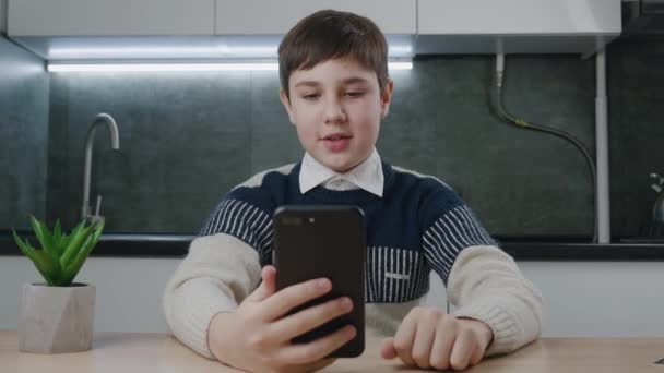 13 anos de idade menino fazendo videochamada usando smartphone enquanto sentado à mesa na cozinha em casa. Adolescente estudante tem um bate-papo de vídeo online na sala de estar, comunicação remota. — Vídeo de Stock