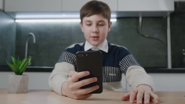 13-летний мальчик делает видеозвонок, используя смартфон, сидя за столом на домашней кухне. Подросток-школьник ведет онлайн-видеочат в гостиной, дистанционное общение. — стоковое видео