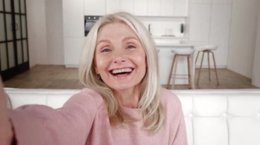 Selfie video konferansında gülümseyen, gülümseyen ve evdeki kanepede oturan neşeli son sınıf öğrencisi. Gülümseyen olgun büyükanne akıllı telefon tutuyor. Evdeki kanepede video görüşmesi yapıyor..