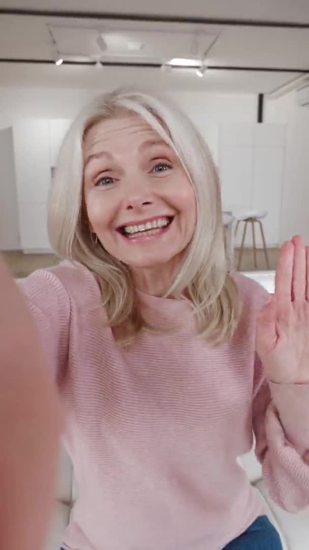 Vídeo vertical de la mujer mayor feliz en la videoconferencia selfie diciendo hola frente a la cámara sonriendo, sentado en el sofá en casa. Sonriente madura abuela sosteniendo teléfono inteligente hacer videollamada en línea — Vídeo de stock