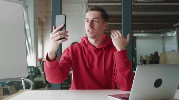 電話画面で人を振って、現代のオフィスでスマートフォンでFacetimeのビデオ通話を行う幸せな男の肖像画。若い男性ブロガー作るvlogビデオブログ使用して携帯電話 — ストック動画