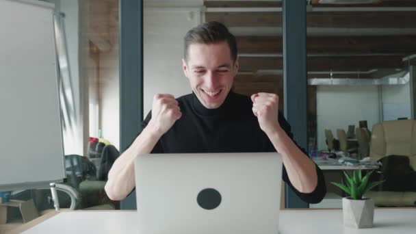 Un om de afaceri de succes stă la masă cu laptop-ul care lucrează la birou. El primește vești bune și este încântat. Câștigătorul fericit de sex masculin sărbătorește succesul și arată da gest — Videoclip de stoc