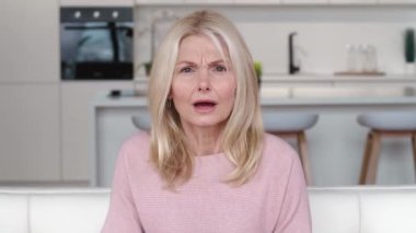 Korkunç haberden korkan son sınıf öğrencisi kadın elleriyle ağzını kapadı ve evdeki kanepede otururken kameraya dehşete kapıldı. İçeride şok olmuş yaşlı bir kadının portresi..