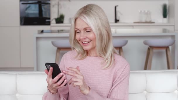 Mujer mayor de pelo gris feliz sosteniendo el teléfono inteligente mirando la pantalla del teléfono móvil jugando juegos móviles utilizando aplicaciones de redes sociales en el teléfono inteligente sentarse en el sofá en casa. — Vídeo de stock