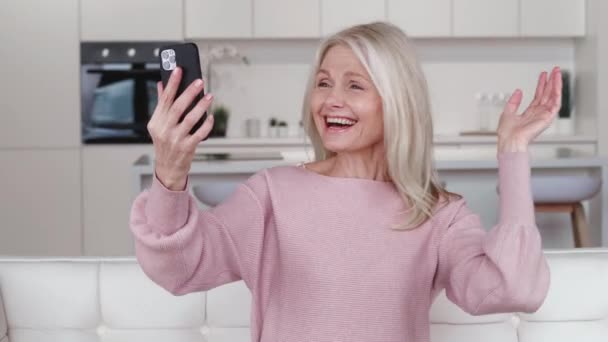 Femme aux cheveux gris joyeux parlant à la vidéoconférence smartphone à la maison, assis sur le canapé. sourire senior mature dame grand-mère parler faire chat vidéo communiquer assis en ligne sur le canapé à la maison. — Video