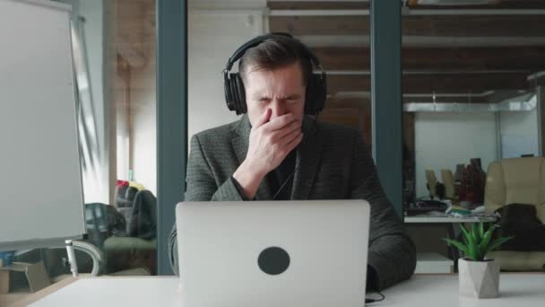 Un jeune homme dans un casque éternuera et se soufflera le nez en travaillant sur un ordinateur portable au bureau. Portrait d'un homme d'affaires malade sur son lieu de travail. — Video