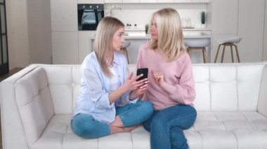 Neşeli beyaz aile portresi, kıdemli anne ve yetişkin kızın gülümsemesi ve birlikte zaman geçirmesi, kanepede oturup akıllı telefondan yazı yazması ve internette alışveriş yapması..